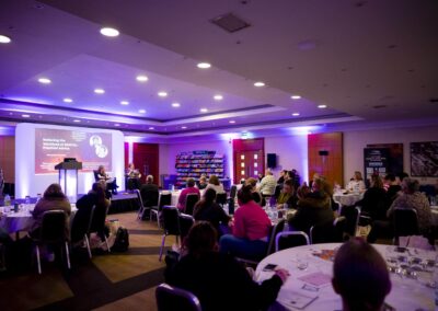 A view from table 8 with Abigail Hawkins FCCT and Lorraine Petersen on stage discussing reducing the workload of SENCOs. Many SENCOs are in the image. A book store sits at the back of the image. At the right hand side you can see Dale Pickles from SENDcast.